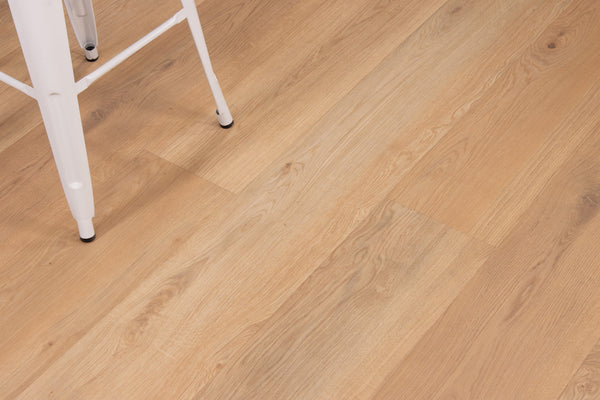 Close-up of light wood flooring with natural grain patterns. The leg of a white metal chair is visible in the top left corner of the image.