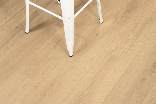 A white metal chair with thin legs sits on light wood grain flooring. The photo focuses on the flooring’s texture and pattern, showing only part of the chair in the upper left corner.