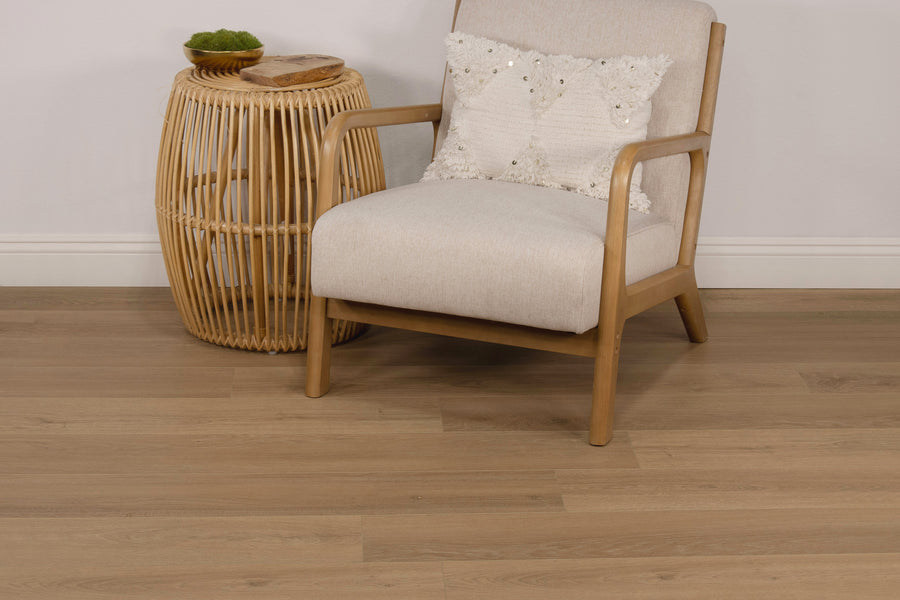 A light-colored wooden armchair with a white textured pillow sits next to a round, wicker side table with a small plant and two wooden coasters on top, on a light wood floor.