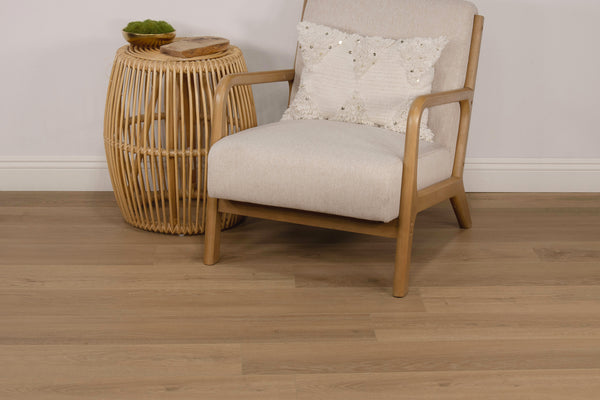 A light-colored wooden armchair with a white textured pillow sits next to a round, wicker side table with a small plant and two wooden coasters on top, on a light wood floor.