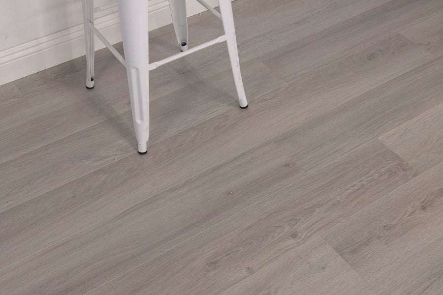 A white metal stool with black-tipped legs on a floor with light gray wood grain planks, next to a white baseboard.