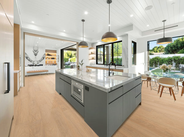 Modern open-concept kitchen with a large gray island, marble countertop, built-in oven, hanging lights, and wood floors. Sliding doors open to a patio with outdoor seating and lush greenery visible outside.