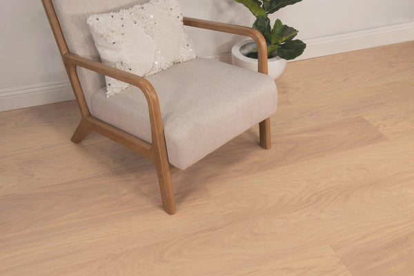 A light wood chair with beige upholstery and a textured white cushion sits on a light wood floor next to a white potted plant against a plain light wall.