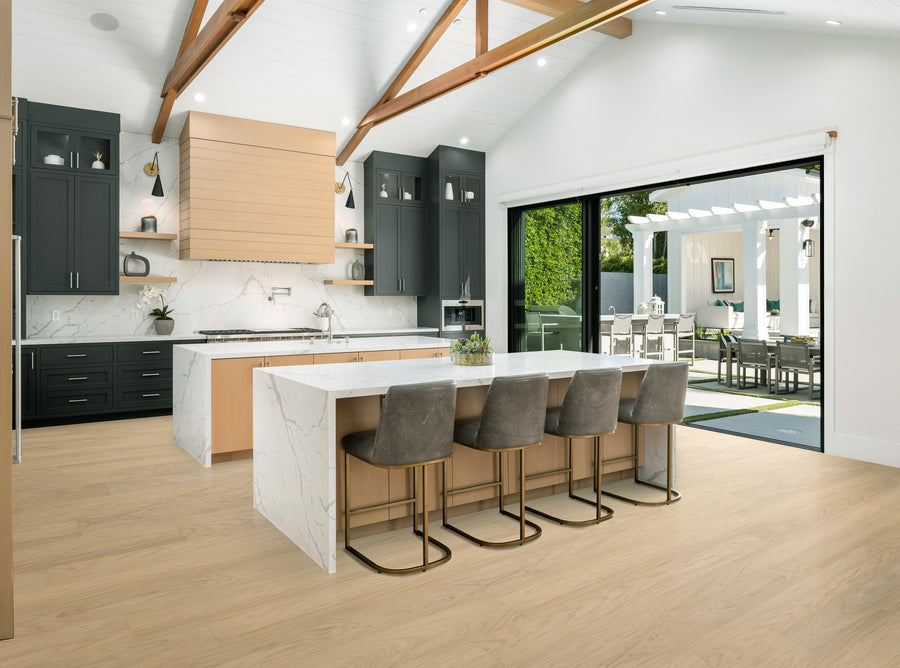 Modern kitchen with light wood floors, a large marble island with four gray barstools, dark cabinets, exposed wooden beams, and sliding glass doors opening to a bright outdoor patio with seating and greenery.