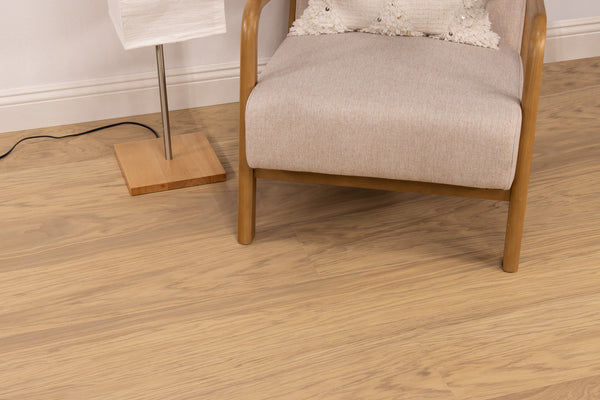 A light wood floor with a beige armchair and a pillow beside a floor lamp with a wooden base and a white shade. The lamps cord is visible on the floor.