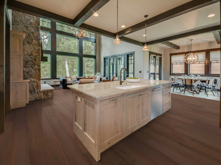 Open, modern kitchen with wooden floors, a large island with sink, pendant lights, and light wood cabinets. Living and dining areas are visible, with large windows letting in natural light and a stone fireplace in the background.