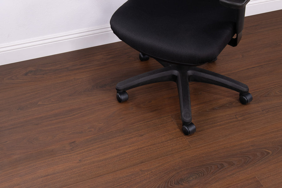 A black office chair with wheels is positioned on a brown wood floor near a white wall with baseboard trim.