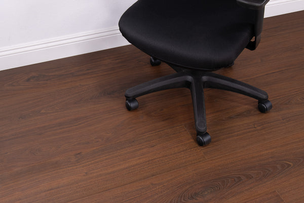 A black office chair with wheels is positioned on a brown wood floor near a white wall with baseboard trim.
