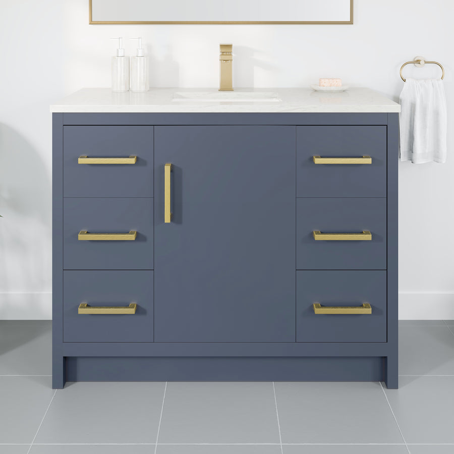 The Curio 42" Vanity w/ Carrara Quartz Top features a navy blue cabinet, gold handles, and an integrated sink. Soap and lotion dispensers are on the left, while a hand towel hangs from a gold ring on the right.