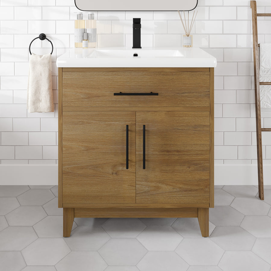 A Graham 30" Transitional Vanity in Oak, featuring black handles and an Arabescato engineered top, sits against a white tiled wall. To the left, a towel hangs from a black ring, while a ladder-style rack stands to the right. Light gray hexagonal floor tiles complement this timeless vanity.
