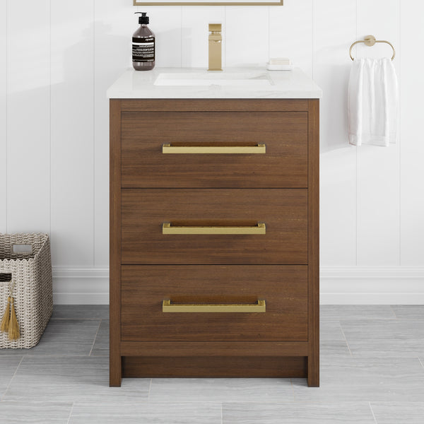 A Curio 24″ Vanity in walnut, featuring three drawers with gold handles and a smooth Carrara quartz top, serves as the focal point of the space. The bathroom vanity is accented by a soap dispenser, a towel ring holding a white towel, and a small basket, all arranged on a gray tiled floor.
