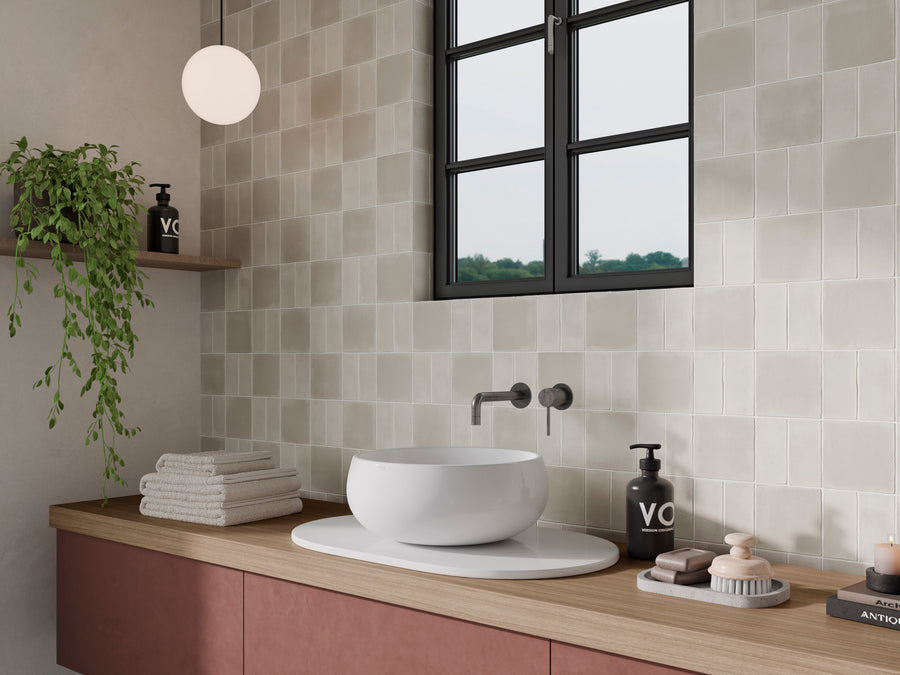 A modern bathroom with a round white sink on wood, wall-mounted faucet, folded towels, soap dispensers, stacked soaps, a potted plant on a shelf, a scenic window view, and unique Harmony 5x5 Porcelain Tile accents.
