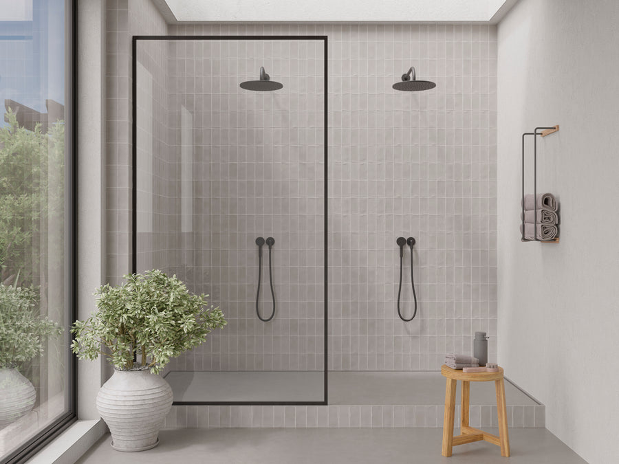 Modern shower with two rain showerheads and hand showers, separated by a glass partition, light gray tile walls, a potted plant, wooden stool with gray towel, and wall shelf with rolled towels and bottles. Large window to the left.