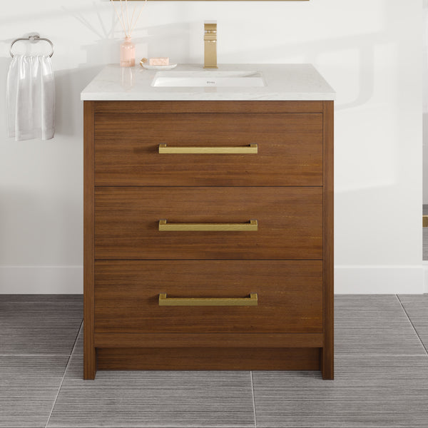 The Curio 30″ Vanity in walnut features three gold-handled drawers, a pristine Carrara quartz top with a white countertop, a rectangular sink, and an elegant gold faucet. A towel hangs on the left wall, while a diffuser and soap dish sit neatly on the counter.