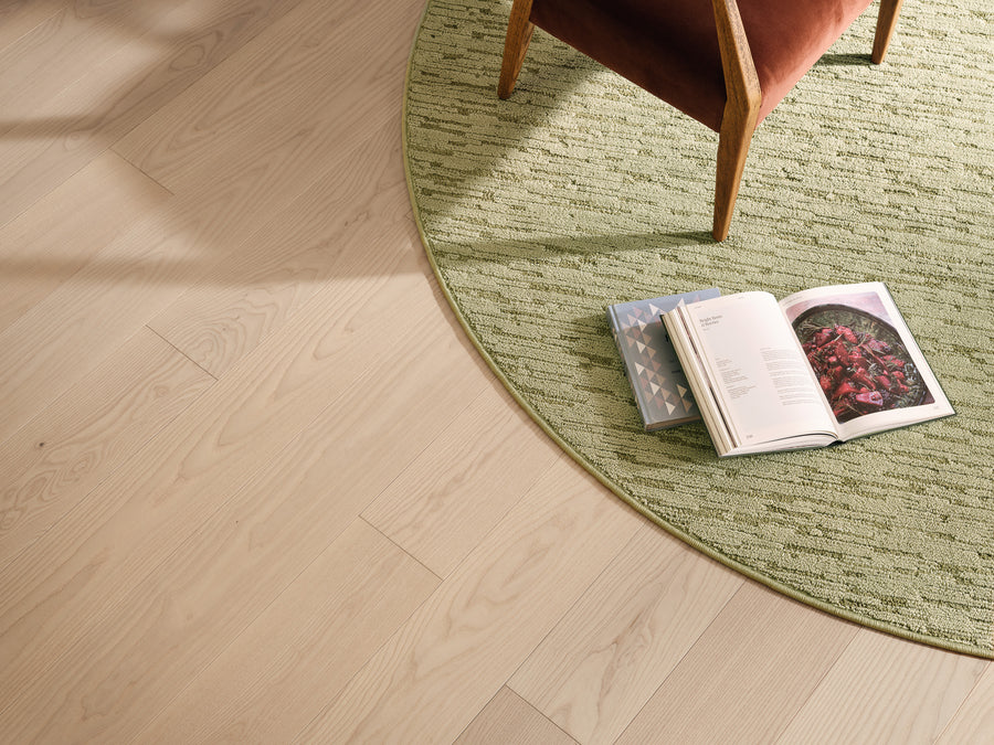 A corner of a wooden floor with a round green rug, a brown chair, an open book showing a photo of red berries, and a closed blue book placed beside it. Sunlight casts soft shadows across the scene.