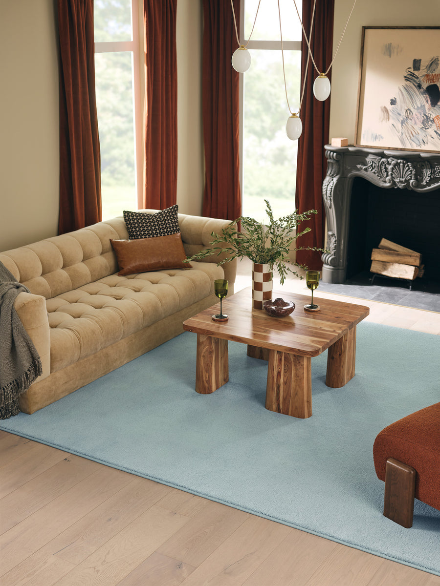 A cozy living room with a beige tufted sofa, a wooden coffee table, two glasses, a vase with greenery, a blue rug, dark red curtains, and a black fireplace with a framed artwork above it.