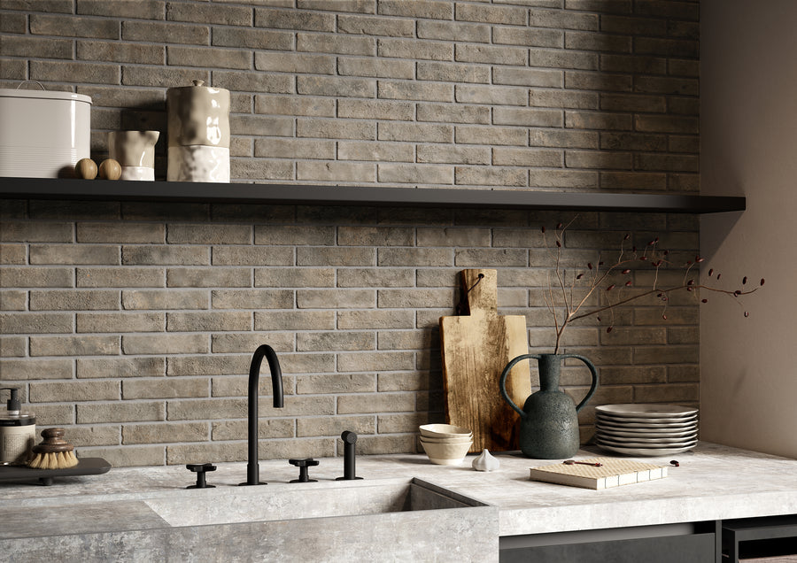 Modern kitchen with gray brick backsplash, black faucet, concrete countertop, and open shelf holding jars and small containers. Counter displays cutting boards, stacked plates, a ceramic jug, and a branch in a vase.