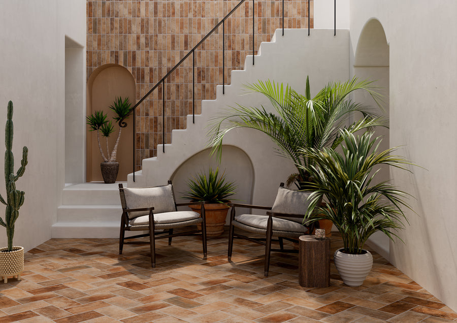A stylish indoor patio with tiled floors and walls, potted plants, two cushioned wooden chairs, a small side table, and a white staircase with a black railing. Natural light brightens the space.