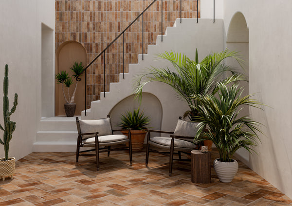 A stylish indoor patio with tiled floors and walls, potted plants, two cushioned wooden chairs, a small side table, and a white staircase with a black railing. Natural light brightens the space.