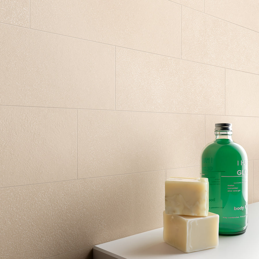 A green bottle, two bars of soap, and a white surface are in the foreground, with a beige tiled wall in the background. The overall scene suggests a clean, minimalist bathroom or spa setting.