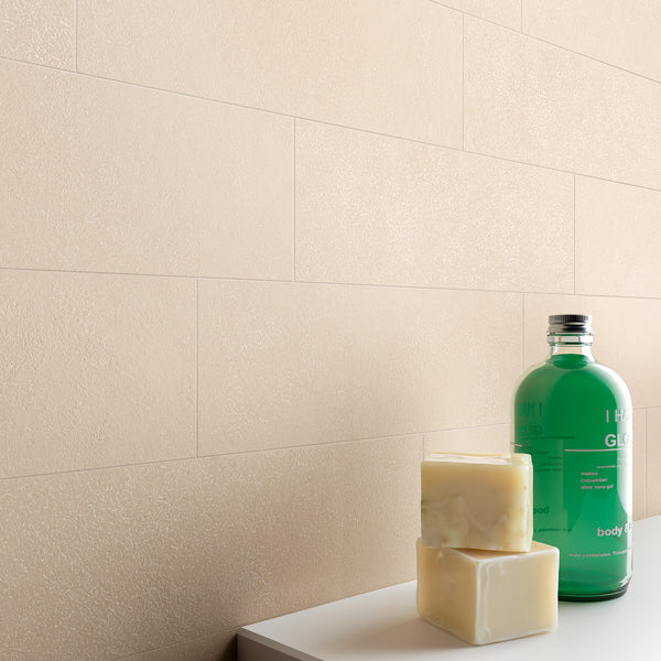 A green bottle, two bars of soap, and a white surface are in the foreground, with a beige tiled wall in the background. The overall scene suggests a clean, minimalist bathroom or spa setting.