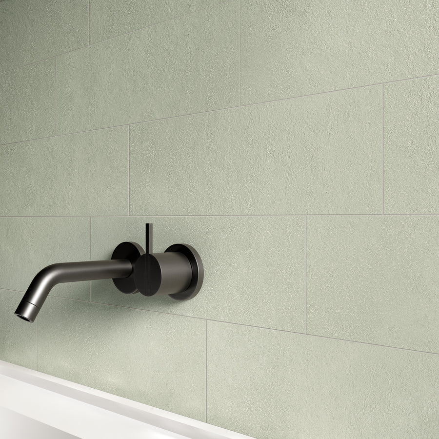 A modern black wall-mounted faucet and handle above a white bathtub, set against a light green textured tile wall.
