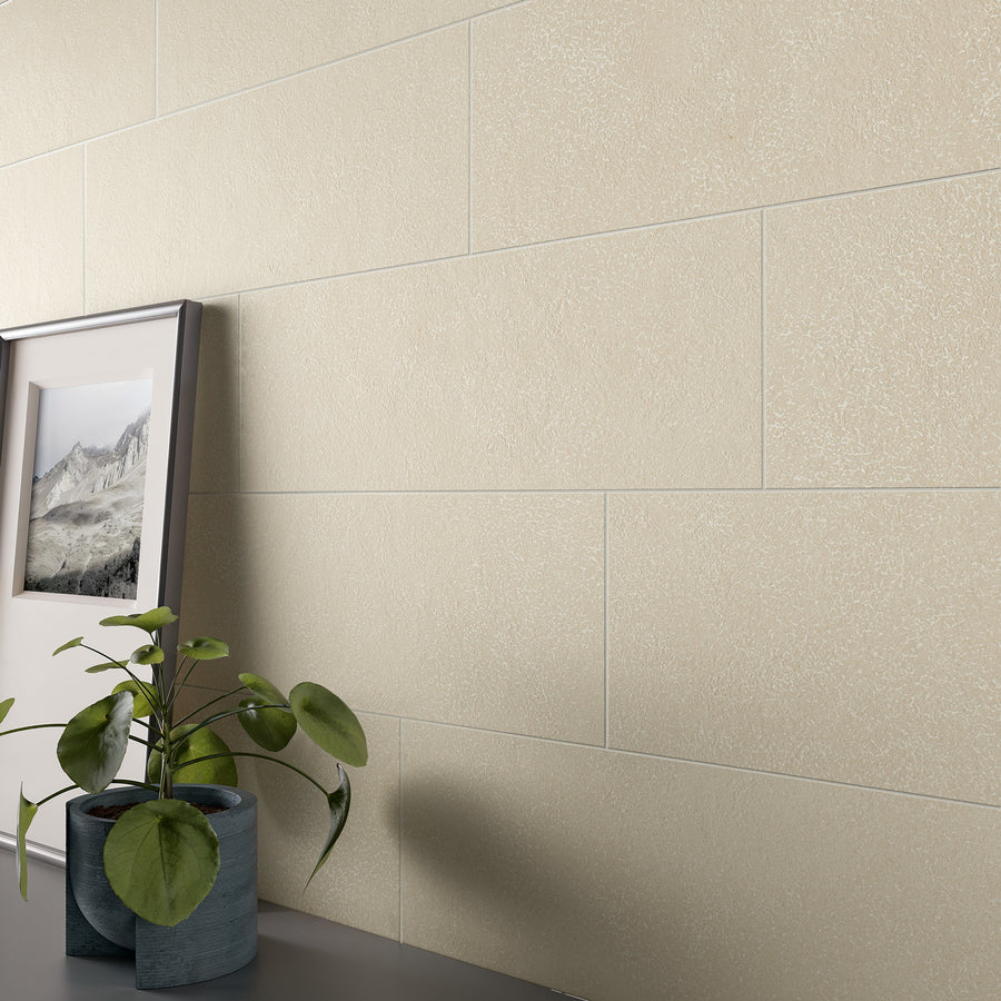 A close-up of a beige tiled wall with a framed landscape photo and a green potted plant on a dark surface in the foreground. The scene appears minimalistic and modern.