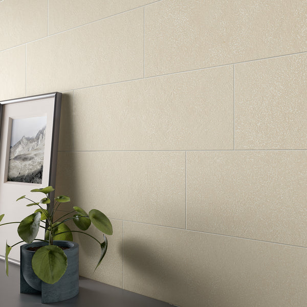 A close-up of a beige tiled wall with a framed landscape photo and a green potted plant on a dark surface in the foreground. The scene appears minimalistic and modern.
