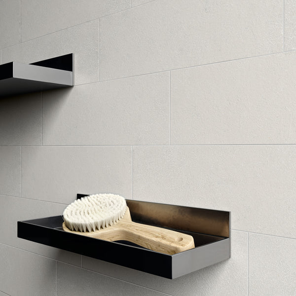 A wooden bath brush with white bristles rests on a sleek, black, wall-mounted shelf against a light beige tiled wall. Another empty shelf is visible above.