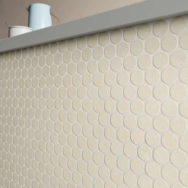 A wall with round, beige textured tiles arranged in a grid pattern. On top of the wall are a blue pitcher and a white container against a plain light-colored background.