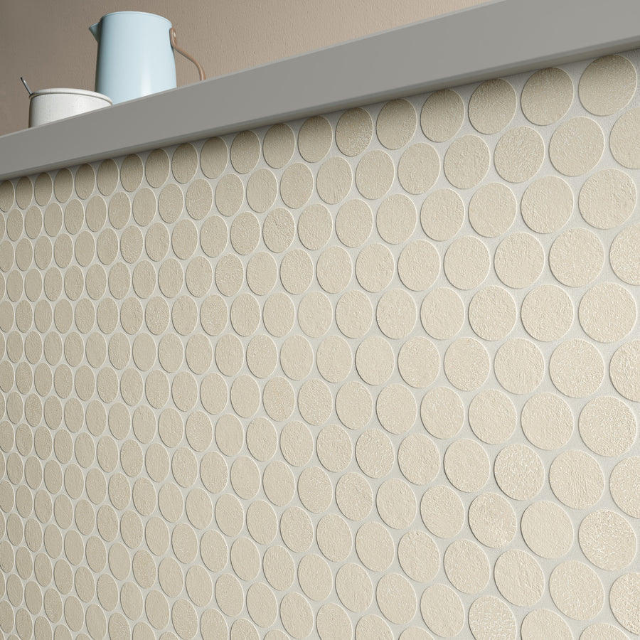 A close-up of a wall covered with round, textured beige tiles. On top of the wall, there is a white bowl and a light blue pitcher. The background is a plain, light-colored wall.