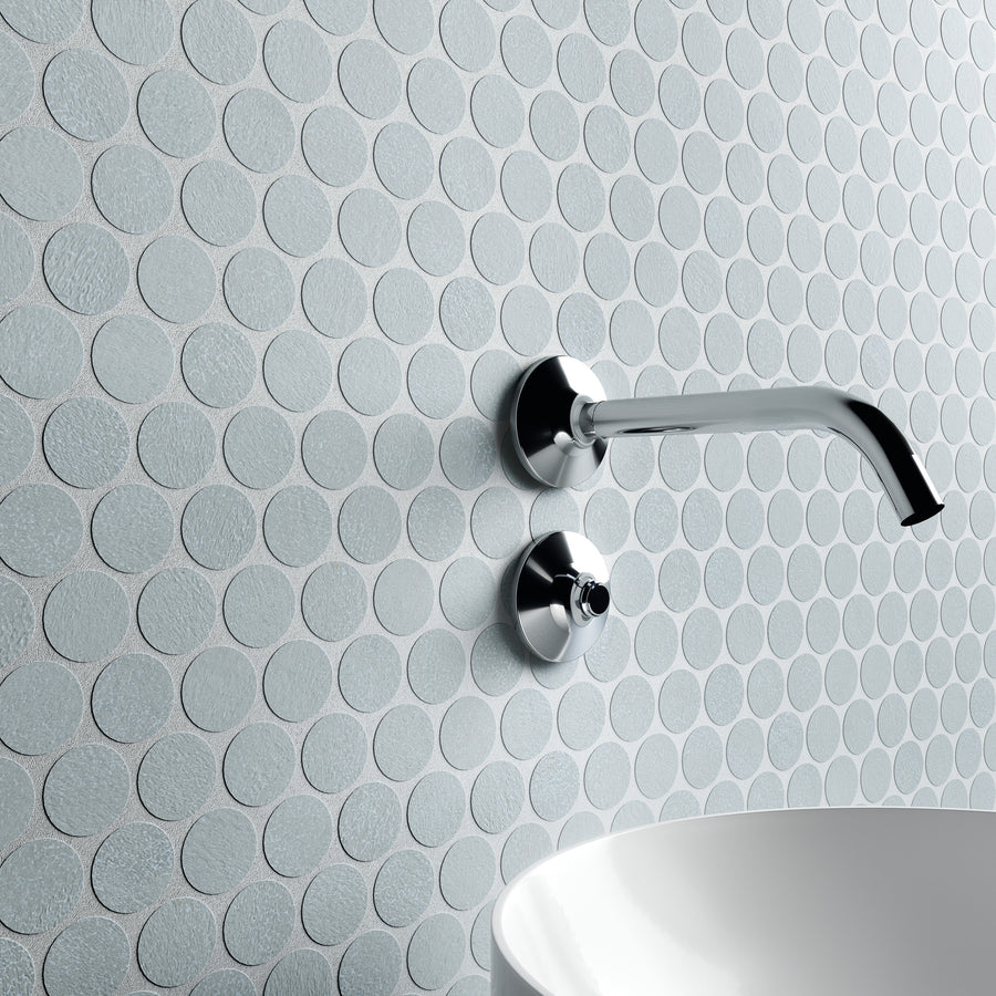 A modern bathroom wall with round, textured, light gray tiles, featuring a minimalist chrome faucet and handle above a white, curved basin sink.