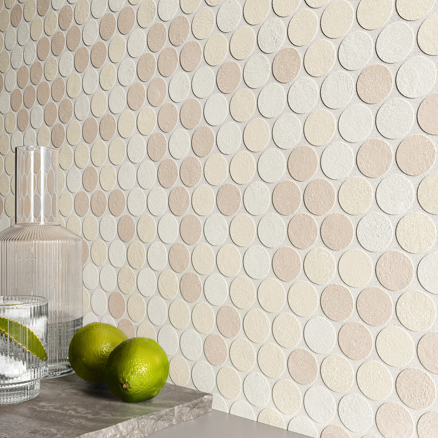 A glass decanter, a glass of water with ice, and two limes sit on a stone surface in front of a beige and white round mosaic tile wall.