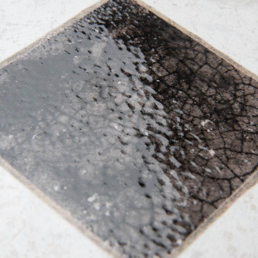 A close-up of a ceramic tile with a glossy finish, featuring a dark, crackled, and textured square pattern in the center, fading slightly at the edges, set against a light background.