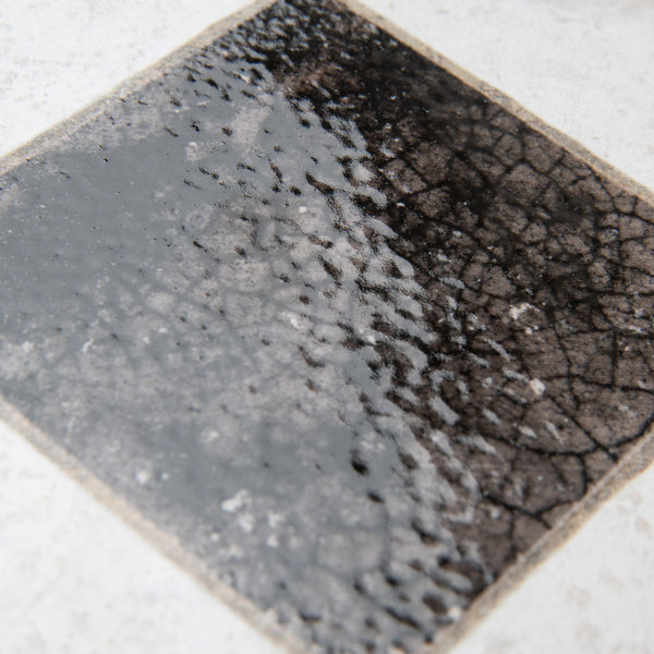 A close-up of a ceramic tile with a glossy finish, featuring a dark, crackled, and textured square pattern in the center, fading slightly at the edges, set against a light background.