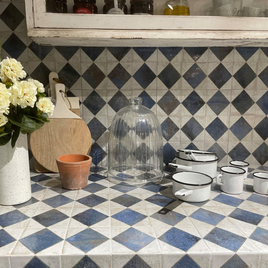 A kitchen counter with blue and white diamond tiles, white enamel pots with black rims, a glass cloche, round wooden cutting boards, a potted plant with white flowers, and an empty terracotta pot.
