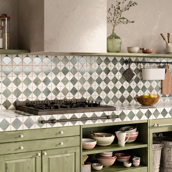 A kitchen with green cabinets, open shelves holding bowls and plates, a gas stove, a tiled backsplash with a diamond pattern, and a countertop with a vase of olive branches and a bowl of fruit.