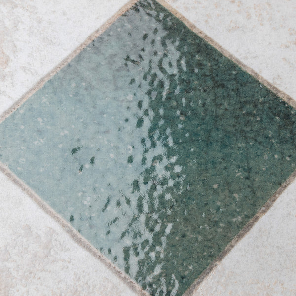 A square-shaped glass tile set diagonally in a light-colored stone surface, with the tile’s translucent greenish hue and textured, rippled surface resembling water.