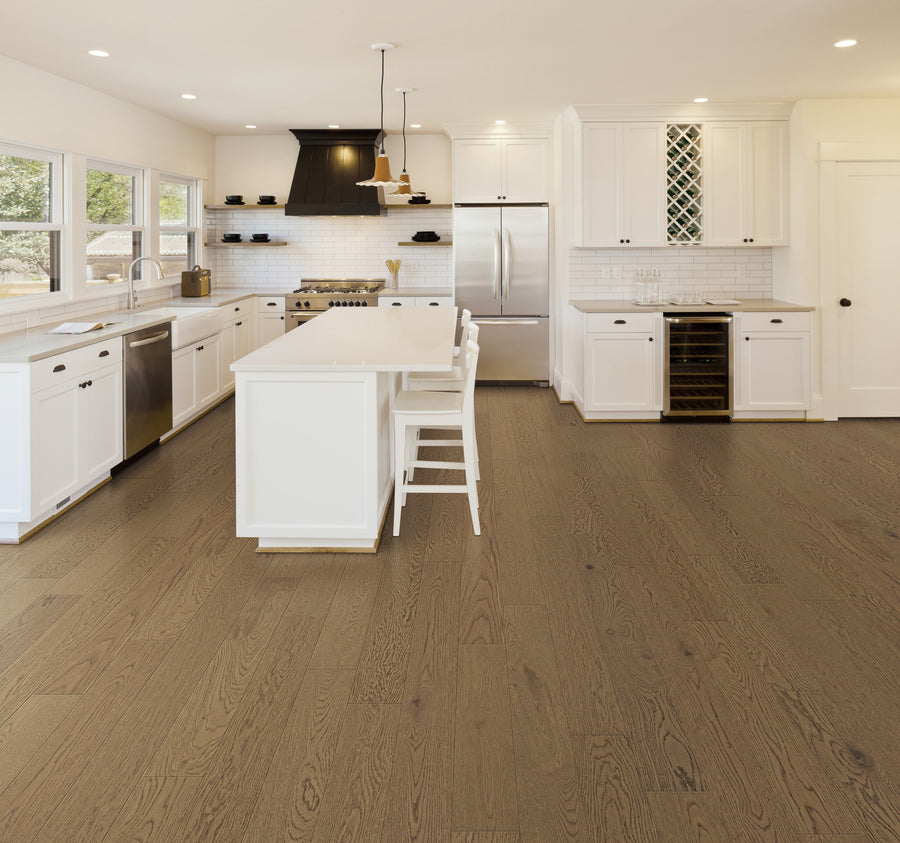 A modern kitchen with white cabinets, stainless steel appliances, a large island with seating, light countertops, hardwood floors, and large windows letting in natural light.