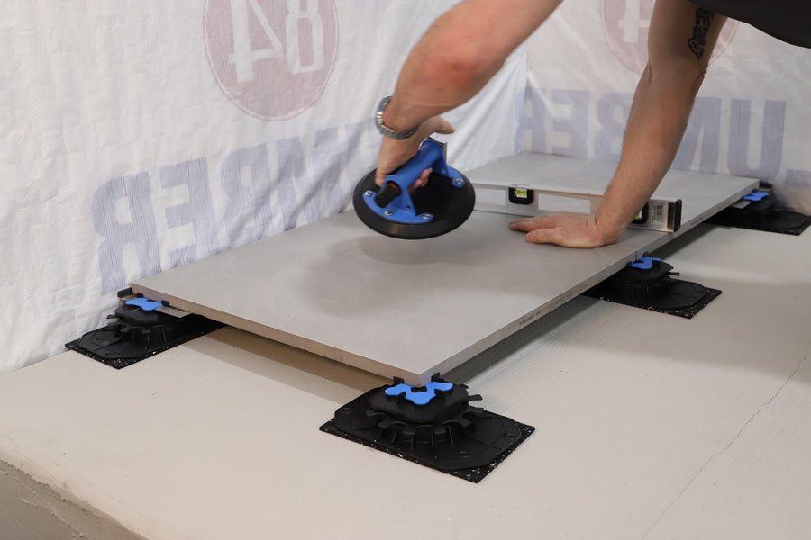 Using a suction cup tool, a person places a large tile onto Ardex TerraMaxx Pedestal supports during a roof terrace install. A level rests on the tile, the smooth base is visible, and the person's tattooed arm can be seen in the shot.