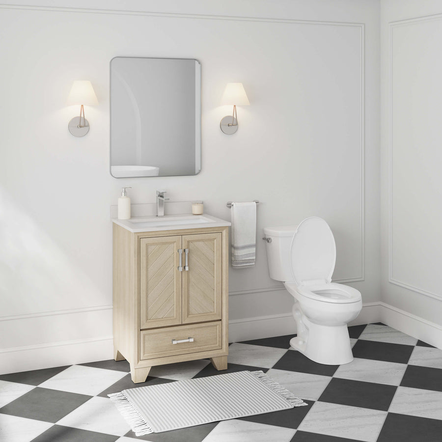 Modern bathroom with a light wood vanity, sink, mirror, and two wall sconces. A white toilet is beside the vanity, and a gray and white checkered floor has a small rug in front of the sink. Walls are light-colored.