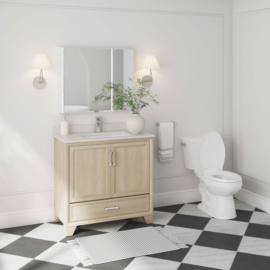 Modern bathroom with a light wood vanity, white countertop, and rectangular mirror. A potted plant, soap dispenser, and towel decorate the space. Black-and-white checkered floor and toilet are also visible.