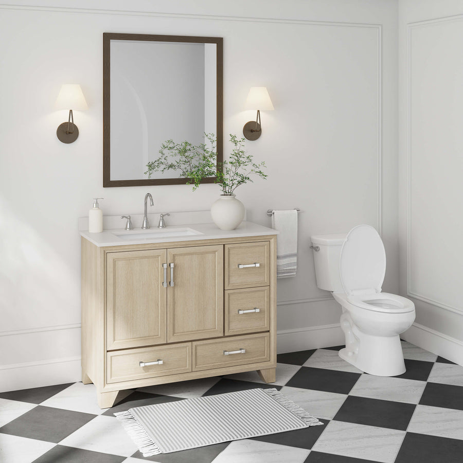 A modern bathroom with a light wood vanity, sink, mirror, and two wall sconces. A potted plant sits on the counter. A white toilet and a towel are on the right, with a rug on black and white checkered tile floor.