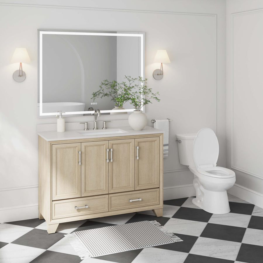 A modern bathroom with a light wood vanity, large mirror, white sink, potted plant, wall sconces, a white toilet, striped rug, and black-and-white checkered floor tiles.