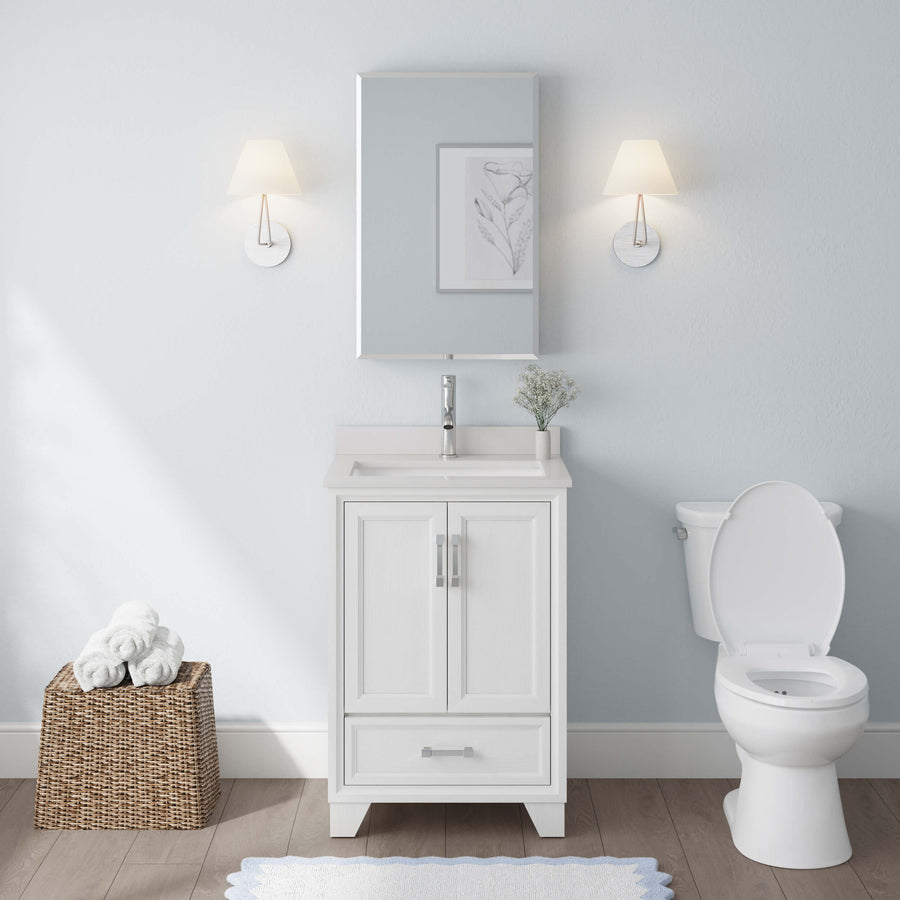 A clean, modern bathroom showcases the Ashlow 24" w/ Engineered Stone Top Vanity with sink and toilet. Above is a mirror flanked by two wall lamps. A wicker basket with rolled towels sits on the floor next to a blue rug.