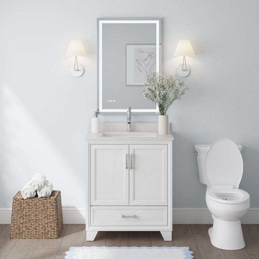 A modern bathroom with the Ashlow 30" w/ Engineered Stone Top Vanity, white sink, rectangular mirror, two wall sconces, toilet, flower vase, and a wicker basket of rolled towels on the wooden floor.