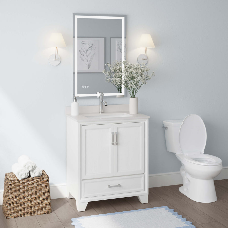 A modern bathroom with a white vanity and sink, a large mirror with lights, a toilet, two wall sconces, framed art, a basket of towels, and a vase with white flowers on the countertop.