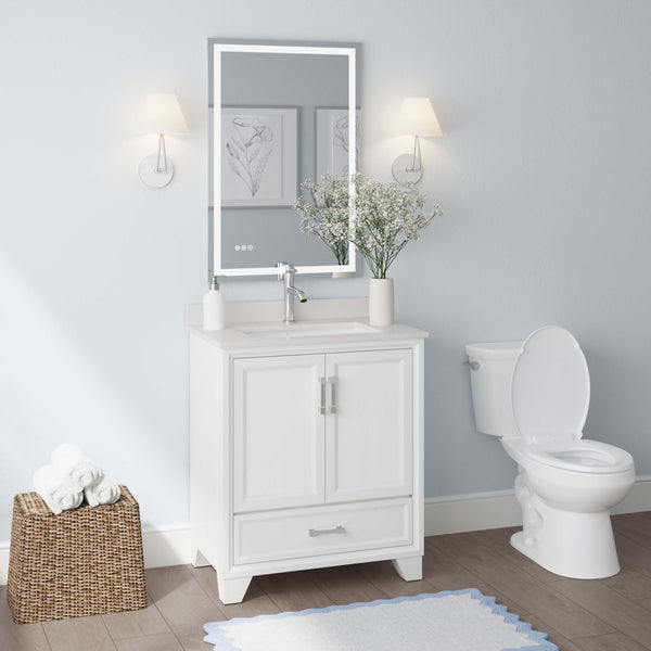 A modern bathroom with a white vanity and sink, a large mirror with lights, a toilet, two wall sconces, framed art, a basket of towels, and a vase with white flowers on the countertop.