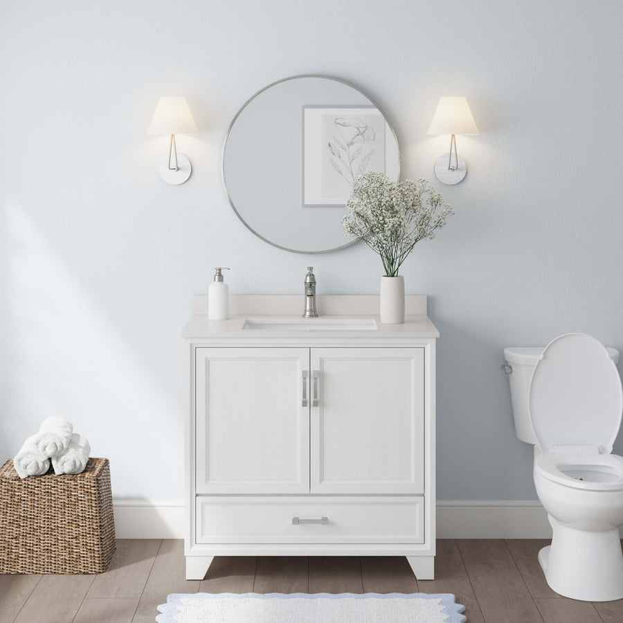 A modern bathroom showcases the Ashlow 36" w/ Engineered Stone Top Vanity, a round mirror, two wall sconces, a flower vase, soap dispenser, toilet, and a wicker basket of white towels on the floor.