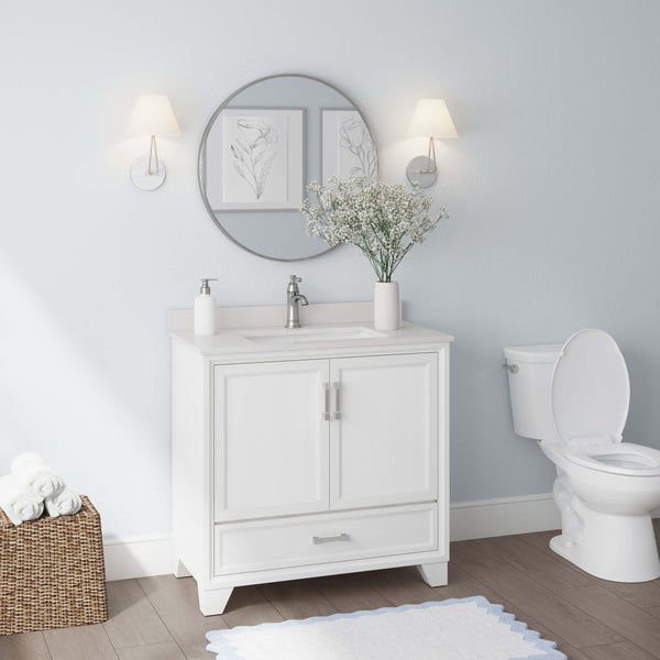 A modern bathroom with a white vanity, round mirror, wall sconces, vase with flowers, toilet, and a basket of rolled towels on a wooden floor with a white rug.